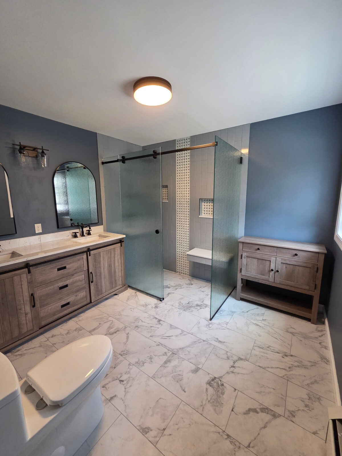 Remodeled bathroom with a double barn-door vanity, arched mirrors, marble-look tile and glass walk-in shower